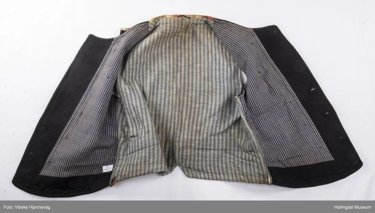 a) åpen mannstrøye, b) mannsvest, c) langbukse, d) halsklut og tilhørende også e) hatt. Open trøye m svarte knappar, dobbelspent vest. Svart m blått, kvitrundut for, rindutt vestebakklut. Sauma maskinsaum til dekor på ermekragar og framside. Ferdig sydd halsklut i silketøy. Filthatt med silkebånd og silkefor.