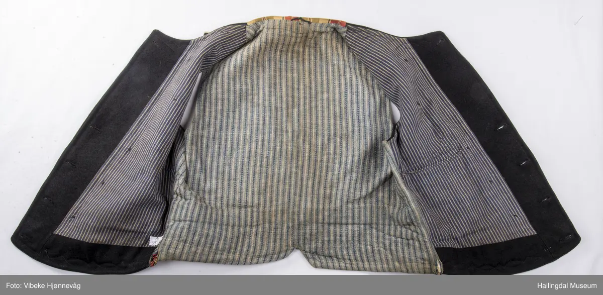 a) åpen mannstrøye, b) mannsvest, c) langbukse, d) halsklut og tilhørende også e) hatt. Open trøye m svarte knappar, dobbelspent vest. Svart m blått, kvitrundut for, rindutt vestebakklut. Sauma maskinsaum til dekor på ermekragar og framside. Ferdig sydd halsklut i silketøy. Filthatt med silkebånd og silkefor.