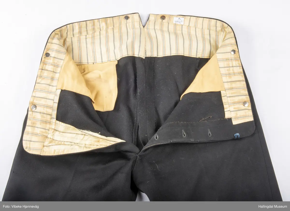 a) åpen mannstrøye, b) mannsvest, c) langbukse, d) halsklut og tilhørende også e) hatt. Open trøye m svarte knappar, dobbelspent vest. Svart m blått, kvitrundut for, rindutt vestebakklut. Sauma maskinsaum til dekor på ermekragar og framside. Ferdig sydd halsklut i silketøy. Filthatt med silkebånd og silkefor.