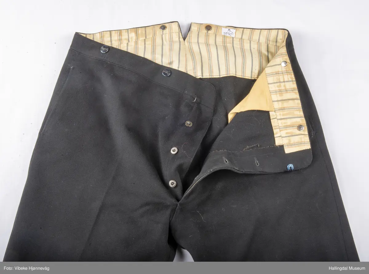 a) åpen mannstrøye, b) mannsvest, c) langbukse, d) halsklut og tilhørende også e) hatt. Open trøye m svarte knappar, dobbelspent vest. Svart m blått, kvitrundut for, rindutt vestebakklut. Sauma maskinsaum til dekor på ermekragar og framside. Ferdig sydd halsklut i silketøy. Filthatt med silkebånd og silkefor.