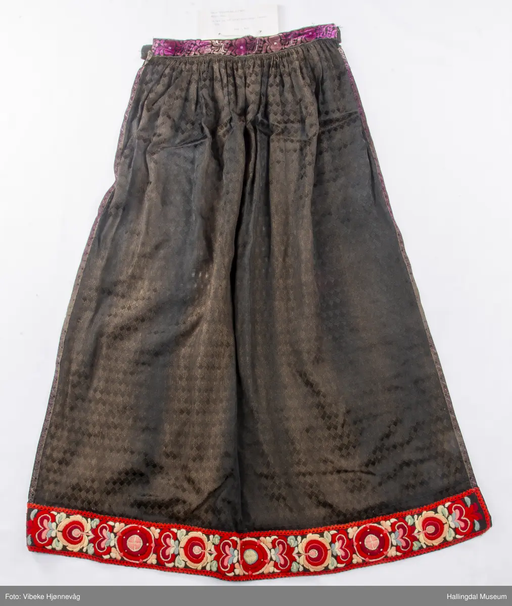 Svart Ulldamask, kroklisser, flanillgådn, eplebånd (fløyel), fløyelsbånd og svart bomullstoff. Lilla og svart bånd, røde kroklisser. Plattsømbroderi: rødt, mørk rødt, rosa, lys rosa, blått, hvitt og lys grønt.
Lengde: 104 cm, bredde: 90 cm, bredde oppe: 41 cm, bredde bånd oppe: 4 cm, bredde plattsømsbroderi nederst: 8,5 cm.