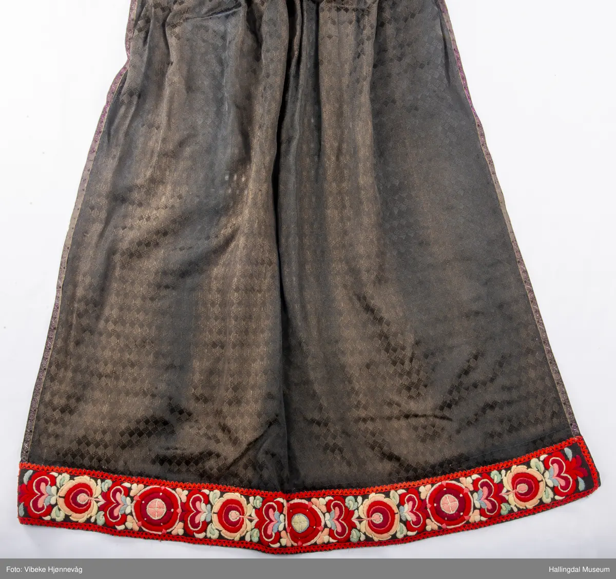 Svart Ulldamask, kroklisser, flanillgådn, eplebånd (fløyel), fløyelsbånd og svart bomullstoff. Lilla og svart bånd, røde kroklisser. Plattsømbroderi: rødt, mørk rødt, rosa, lys rosa, blått, hvitt og lys grønt.
Lengde: 104 cm, bredde: 90 cm, bredde oppe: 41 cm, bredde bånd oppe: 4 cm, bredde plattsømsbroderi nederst: 8,5 cm.