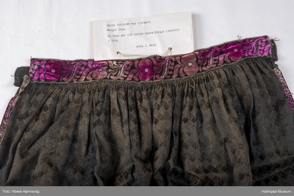 Svart Ulldamask, kroklisser, flanillgådn, eplebånd (fløyel), fløyelsbånd og svart bomullstoff. Lilla og svart bånd, røde kroklisser. Plattsømbroderi: rødt, mørk rødt, rosa, lys rosa, blått, hvitt og lys grønt.
Lengde: 104 cm, bredde: 90 cm, bredde oppe: 41 cm, bredde bånd oppe: 4 cm, bredde plattsømsbroderi nederst: 8,5 cm.