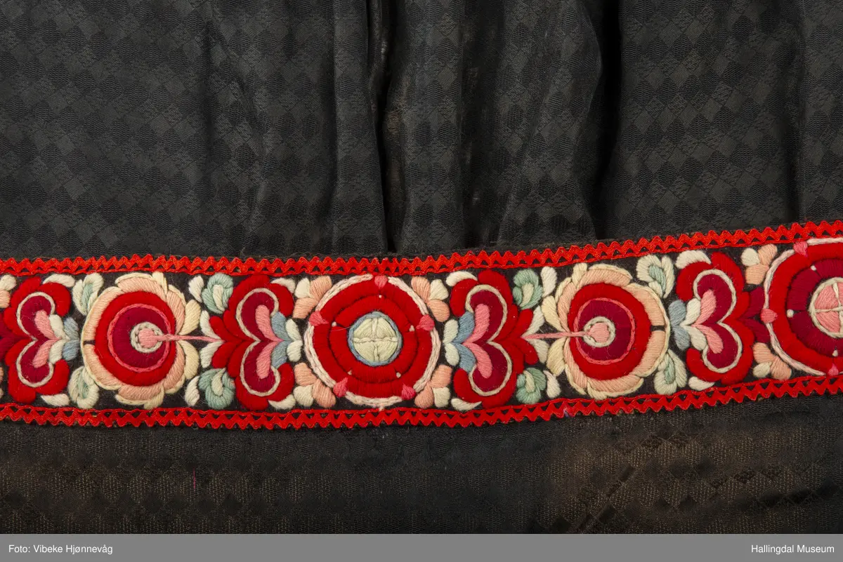 Svart Ulldamask, kroklisser, flanillgådn, eplebånd (fløyel), fløyelsbånd og svart bomullstoff. Lilla og svart bånd, røde kroklisser. Plattsømbroderi: rødt, mørk rødt, rosa, lys rosa, blått, hvitt og lys grønt.
Lengde: 104 cm, bredde: 90 cm, bredde oppe: 41 cm, bredde bånd oppe: 4 cm, bredde plattsømsbroderi nederst: 8,5 cm.