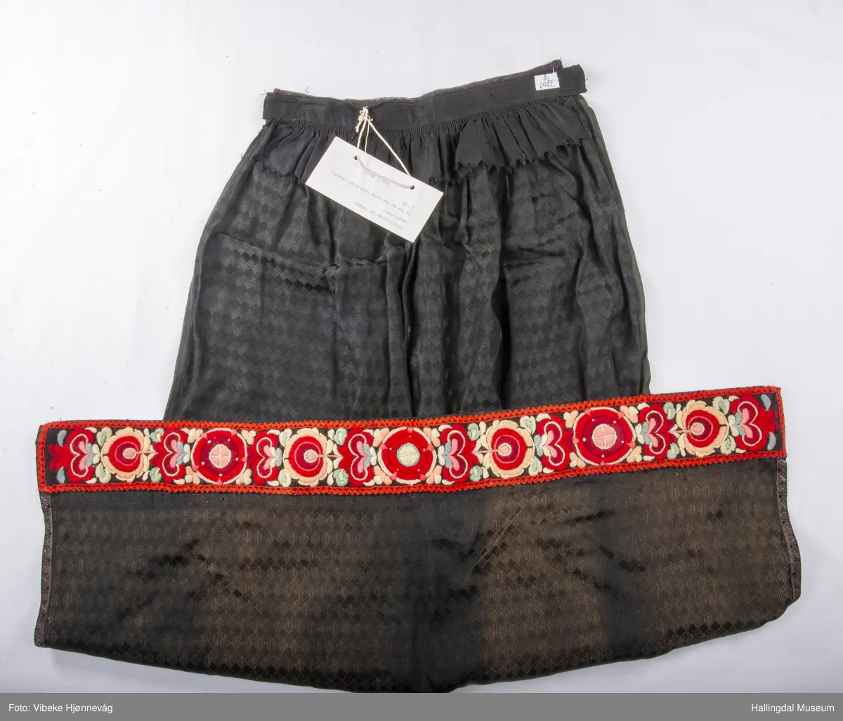 Svart Ulldamask, kroklisser, flanillgådn, eplebånd (fløyel), fløyelsbånd og svart bomullstoff. Lilla og svart bånd, røde kroklisser. Plattsømbroderi: rødt, mørk rødt, rosa, lys rosa, blått, hvitt og lys grønt.
Lengde: 104 cm, bredde: 90 cm, bredde oppe: 41 cm, bredde bånd oppe: 4 cm, bredde plattsømsbroderi nederst: 8,5 cm.