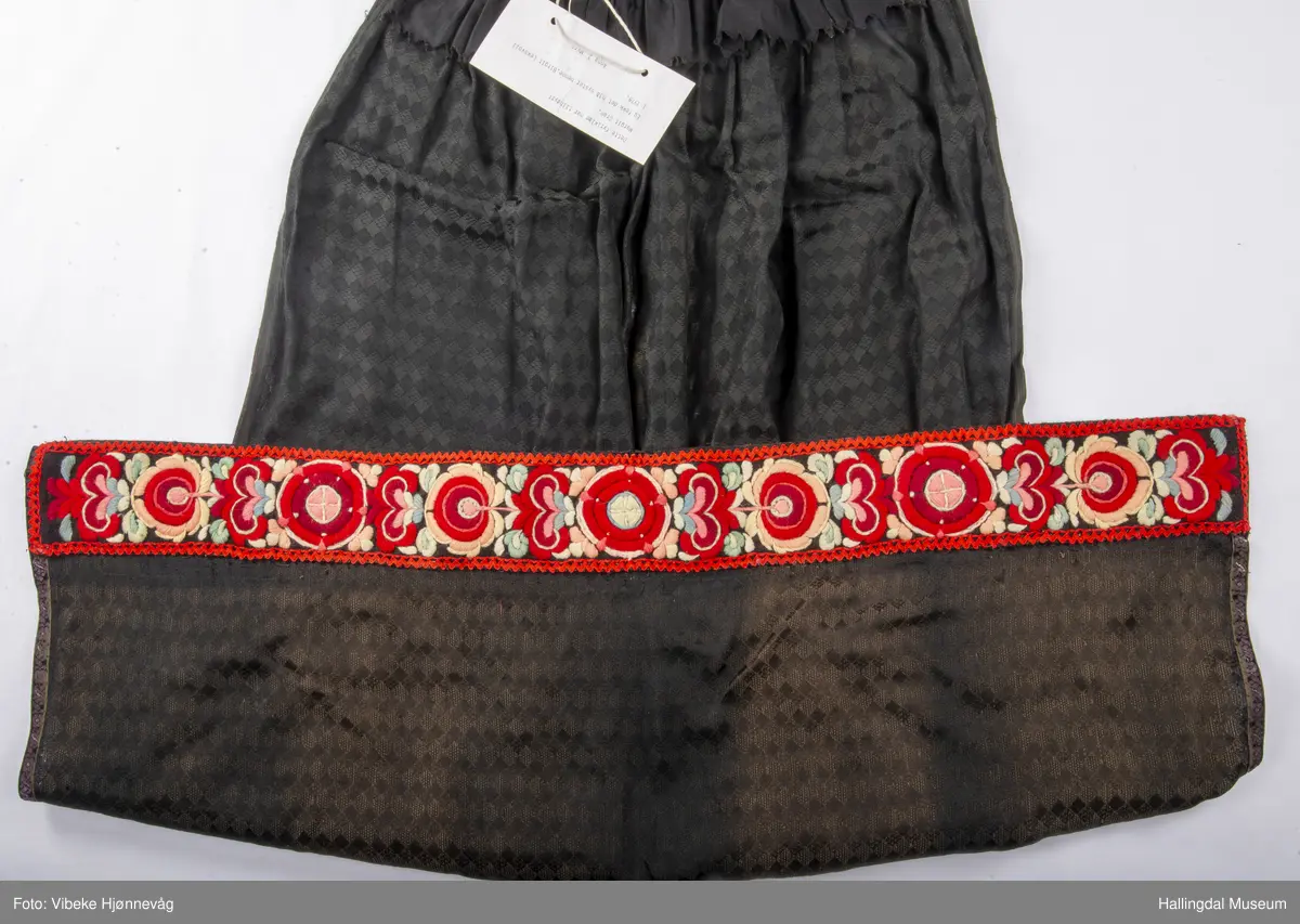 Svart Ulldamask, kroklisser, flanillgådn, eplebånd (fløyel), fløyelsbånd og svart bomullstoff. Lilla og svart bånd, røde kroklisser. Plattsømbroderi: rødt, mørk rødt, rosa, lys rosa, blått, hvitt og lys grønt.
Lengde: 104 cm, bredde: 90 cm, bredde oppe: 41 cm, bredde bånd oppe: 4 cm, bredde plattsømsbroderi nederst: 8,5 cm.