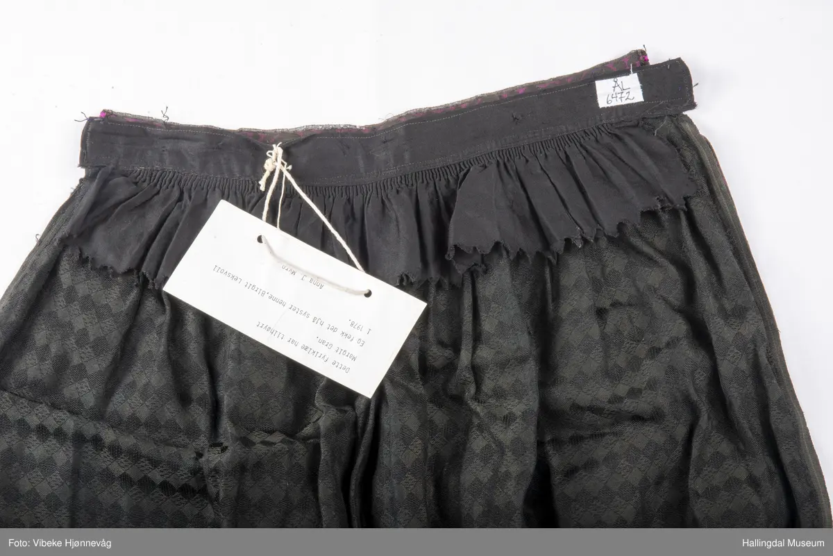 Svart Ulldamask, kroklisser, flanillgådn, eplebånd (fløyel), fløyelsbånd og svart bomullstoff. Lilla og svart bånd, røde kroklisser. Plattsømbroderi: rødt, mørk rødt, rosa, lys rosa, blått, hvitt og lys grønt.
Lengde: 104 cm, bredde: 90 cm, bredde oppe: 41 cm, bredde bånd oppe: 4 cm, bredde plattsømsbroderi nederst: 8,5 cm.