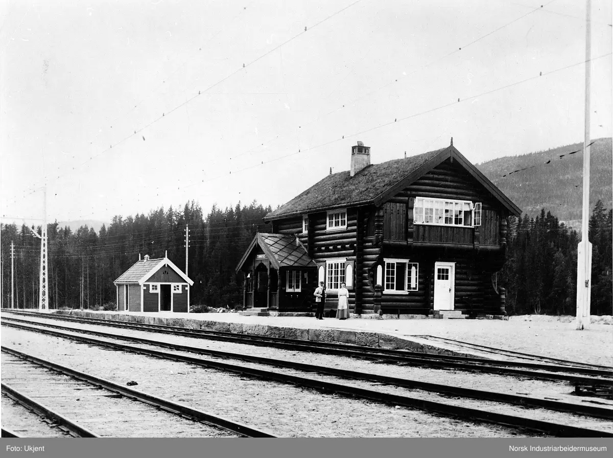 Tinnoset stasjon. To mennesker stående utenfor laftet stasjonsbygning. Flaggstang og uthus stående på perrongen. Tre jernbanespor foran perrongen. Strømmaster langs sporet.