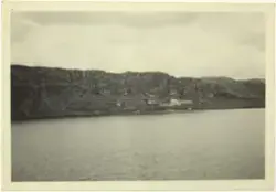 Ein gard ved eit vatn med fjellknausar rett bakom. På baksid