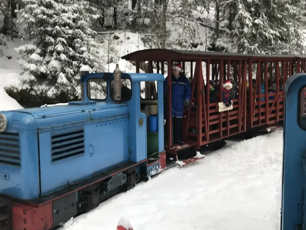 Et blått lite lokomotiv, med røde vogner med barn i, som kjører gjennom snødekket skog.