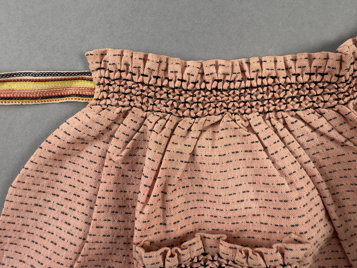 Rosa förkläde, handvävt i tuskaft med inslag av bruna "hopprosor" i bomullgarn. Förklädet har två utanpåhängande *fickor med smocksöm i övre kant. Förklädets midjelinje har också smocksöm sydd med svart tråd i 3mm glesa rader. Traskanten upptill är nervikt mot avigsidan och dolt av ett handvävt band i varprips av bomullsgarn. Bandet är fastsytt med smygstygn runt om samt fortsätter som knytband 62 och 64cm ut på var sida. Långsidorna och nederkant har 3mm respektive 17mm bred dubbelvikt fåll, som är fastsydd med smygstygn. 
* Fickdelen (ca 15x23,5cm) lagd räta mot räta på förklädet är fastsytt med en raksöm ca 8cm lång längs ena fickdelens kortsida. Kortsidans traskant, gömd i en 8mm bred dubbelvikt fåll, vikt mot fickans insida är sydd med smygstygn. Den andra kortsidan är fållad på samma sätt innan den sytts med smocksöm i 5mm glesa rader. Därefter är fickdelen vikt (botten) räta mot räta och ihopsydd med raksöm längs långsidorna, men fickans utsida med smocksöm är lagd 10mm ovanför för att dölja den andra kortsidans fåll. Denna del, 10mm, har sytts fast och fållats på insidan med smygstygn. Se bilder 5-11 på fickan.