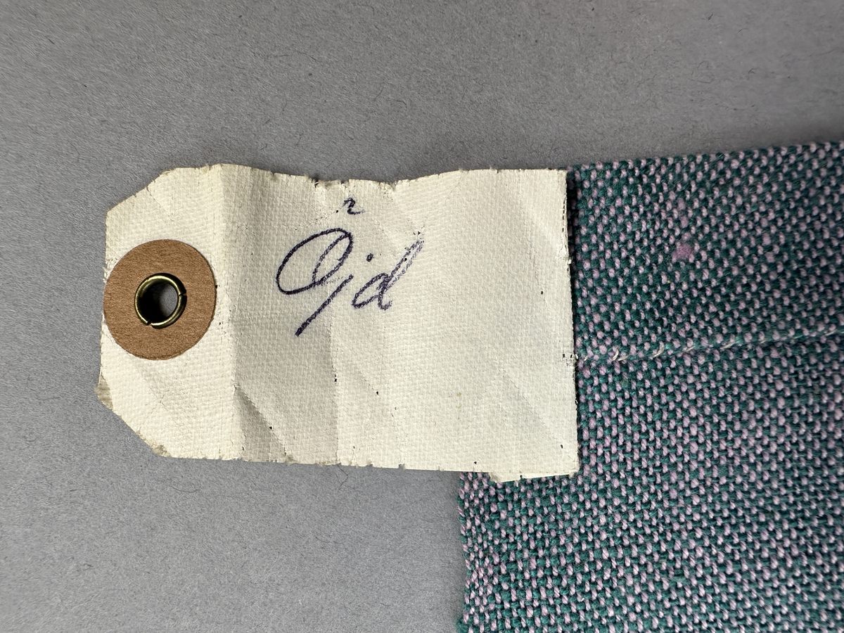 Rutigt förkläde, handvävt i tuskaft och med munkabältesvariation av bomullsgarn. Förklädet har tre inåt lagda veck 1,8cm djupa med 3cm mellanrum på var sida om mitten. Linningen, gjord av dubbla handvävda band i varprips av bomullsgarn, är fastsydda med smygstygn längs tre sidor och upptill med kaststygn. Framsidans band fortsätter som knytband 62 och 63cm på var sida. Nertill har förklädet en dubbelvikt fåll 2,2cm bred sydd med smygstygn och kortsidorna med kaststygn. På högra sidan finns en utanpå ficka, som är fastsydd med 2 parallella raksömmar. Upptill har fickan en dubbelvikt fåll 1,3cm bred sydd med smygstygn.