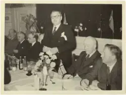 Sju menn ved eit festdekka bord. Ein av dei står og held ein