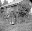 Hulda Svensdotter framför bostaden Näsastugan i Asby. Hennes stuga var enkel men omgivningarna vackra intill Rankhallssjön. När fotografen passerade 1937 var hon nära 80 år och hon skulle komma att leva på platsen till nära 90. Källorna beskriver ett hårt liv, ett typiskt för fattigsverige. Född i Rumskulla dagen före julafton 1858 tycks hennes tillvaro startat med goda förutsättningar. Fadern var rättare på gården Norrhult men något skedde i hast. I ett kommande steg tjänade han som statardräng i Lofta men lyfte sig något för att bli torpare i Sund socken. Hulda lämnade hemmet i 20-årsåldern för ett slitsamt liv som tjänstehjon och piga på gårdar i Ydre med omnejd. Utanför äktenskap födde hon två söner och det bidrog sannolikt till att hon från år 1895 blev erbjuden Näsastugan som inhysehjon.