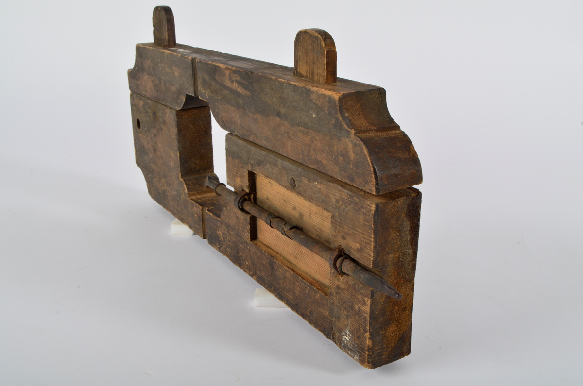 Furu. Snekret. Tilnærmet rektangulær form, innskjæringer ved tre av hjørnene. Redskapet består av to trestykker føyd sammen ved hjelp av to eiketapper som er avrundet i endene. Det ene stykket har kunnet gli på tappene og har antagelig fungert som klemme. Midt på redskapet, stor, kvadratisk åpning med konvekse buer i hjørnene. Over dette, rektangulær nedsenkning, på langs av denne er festet jernbor ved hjelp av to jernbøyler, festet på baksiden med skruegjenger og muttere. Tverrgående hulkil på hver side av den kvadratiske åpningen.