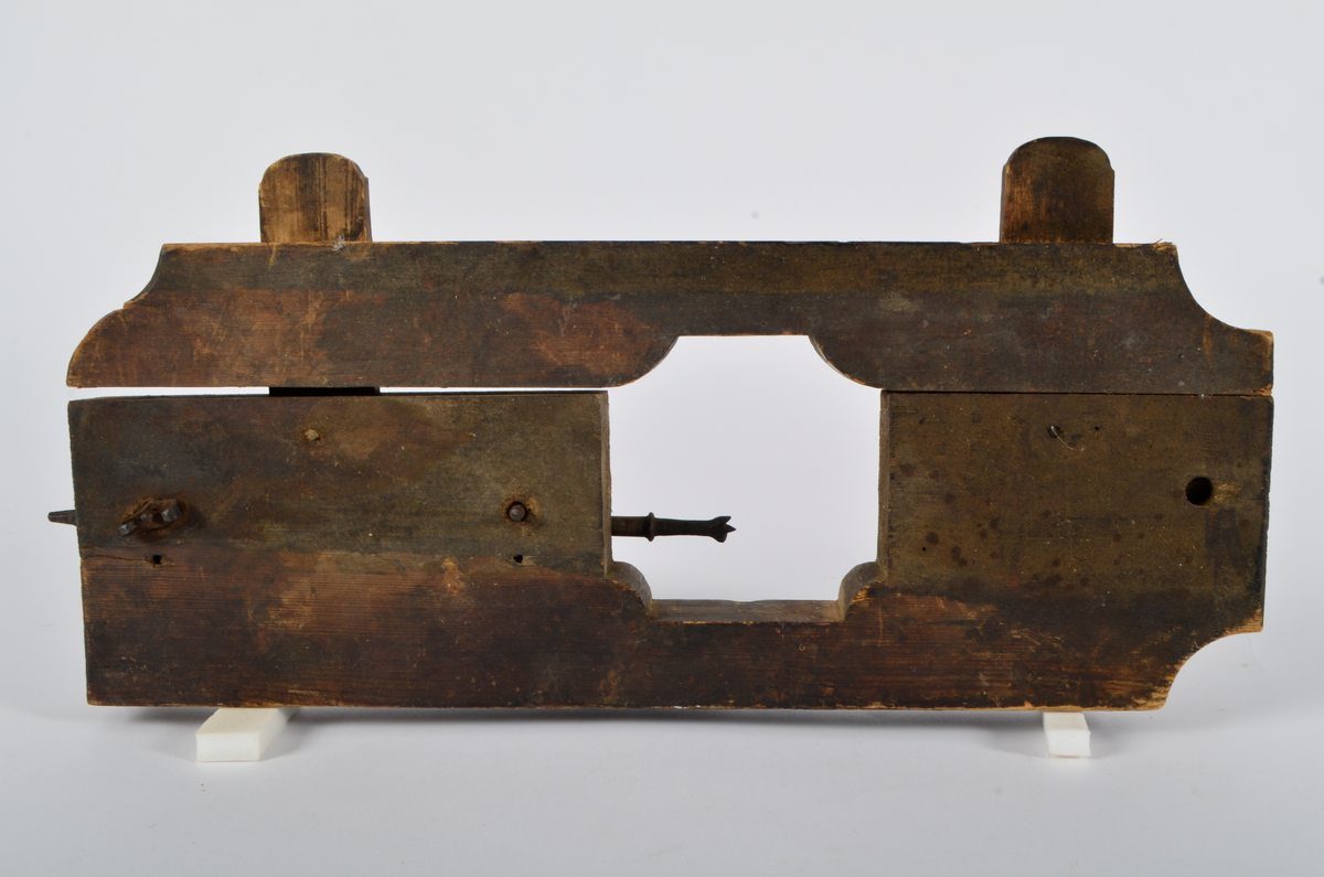 Furu. Snekret. Tilnærmet rektangulær form, innskjæringer ved tre av hjørnene. Redskapet består av to trestykker føyd sammen ved hjelp av to eiketapper som er avrundet i endene. Det ene stykket har kunnet gli på tappene og har antagelig fungert som klemme. Midt på redskapet, stor, kvadratisk åpning med konvekse buer i hjørnene. Over dette, rektangulær nedsenkning, på langs av denne er festet jernbor ved hjelp av to jernbøyler, festet på baksiden med skruegjenger og muttere. Tverrgående hulkil på hver side av den kvadratiske åpningen.