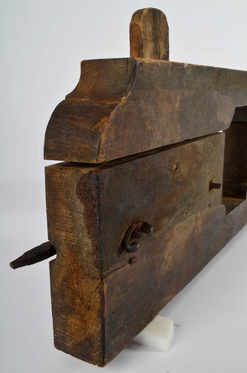 Furu. Snekret. Tilnærmet rektangulær form, innskjæringer ved tre av hjørnene. Redskapet består av to trestykker føyd sammen ved hjelp av to eiketapper som er avrundet i endene. Det ene stykket har kunnet gli på tappene og har antagelig fungert som klemme. Midt på redskapet, stor, kvadratisk åpning med konvekse buer i hjørnene. Over dette, rektangulær nedsenkning, på langs av denne er festet jernbor ved hjelp av to jernbøyler, festet på baksiden med skruegjenger og muttere. Tverrgående hulkil på hver side av den kvadratiske åpningen.