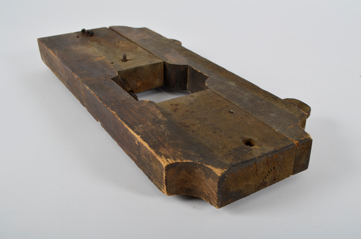 Furu. Snekret. Tilnærmet rektangulær form, innskjæringer ved tre av hjørnene. Redskapet består av to trestykker føyd sammen ved hjelp av to eiketapper som er avrundet i endene. Det ene stykket har kunnet gli på tappene og har antagelig fungert som klemme. Midt på redskapet, stor, kvadratisk åpning med konvekse buer i hjørnene. Over dette, rektangulær nedsenkning, på langs av denne er festet jernbor ved hjelp av to jernbøyler, festet på baksiden med skruegjenger og muttere. Tverrgående hulkil på hver side av den kvadratiske åpningen.