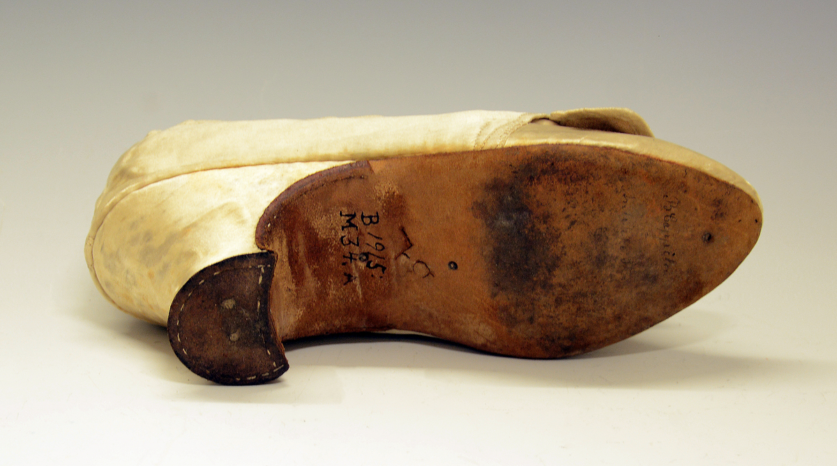 Sko. Silkesko med lærsåler og hæl trukket med gulhvit silke. Avrundet tå og to bånd av samme tøy som ligger i kryss foran. Silkeskoene tilhører drakten  TGM-BM.1914:143
