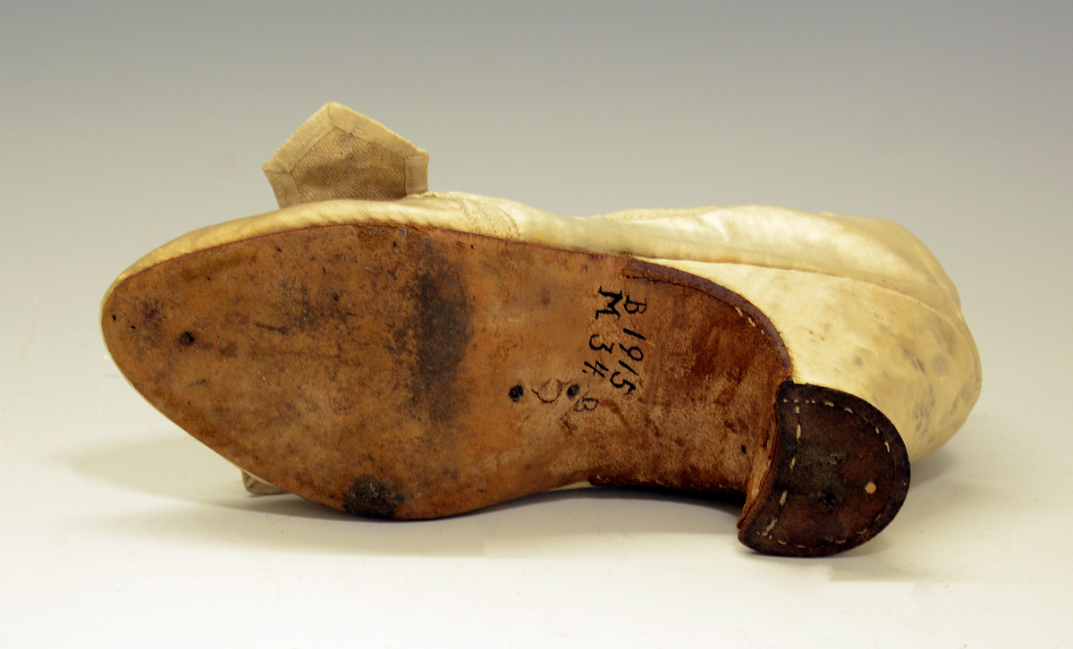 Sko. Silkesko med lærsåler og hæl trukket med gulhvit silke. Avrundet tå og to bånd av samme tøy som ligger i kryss foran. Silkeskoene tilhører drakten  TGM-BM.1914:143