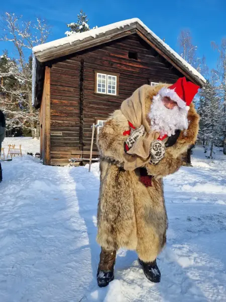 Julenisse med sekk på ryggen i stor ulveskinnspels, foran tømmerhuset Engumstua på Eiktunet museum, Gjøvik. Snø og blå himmel.