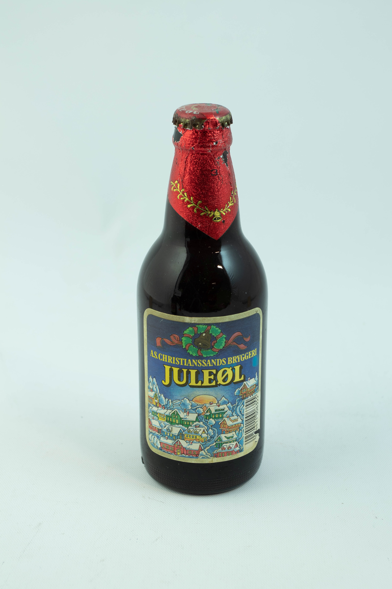 Brun glassflaske med flerfarget etikett med julemotiv. En snødekt landsby.