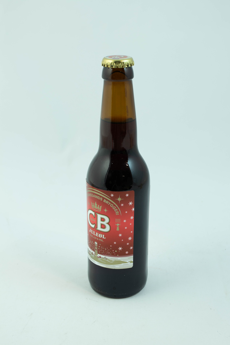 Brun glassflaske med flerfarget etikett med julemotiv. Gyllent fyrtån med hvite snødetaljer på rød bakgrunn