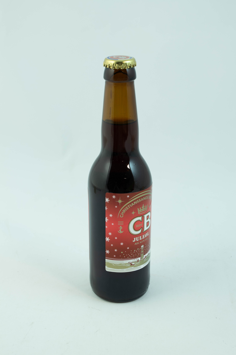 Brun glassflaske med flerfarget etikett med julemotiv. Gyllent fyrtån med hvite snødetaljer på rød bakgrunn