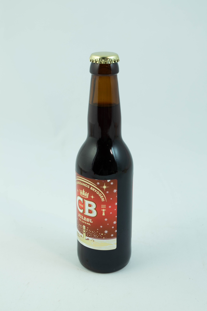 Brun glassflaske med flerfarget etikett med julemotiv. Skinnende gyllent fyrtån med hvite snødetaljer på rød bakgrunn