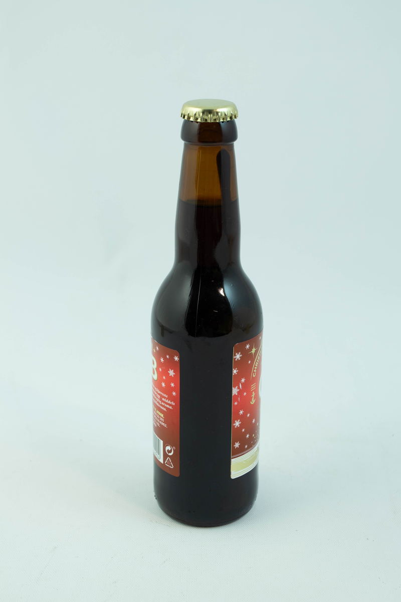 Brun glassflaske med flerfarget etikett med julemotiv. Skinnende gyllent fyrtån med hvite snødetaljer på rød bakgrunn