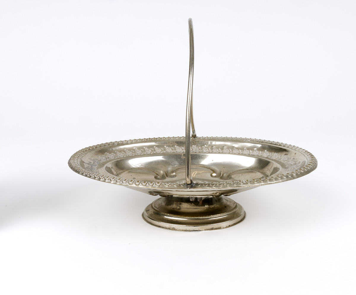 Skål. Oval og flat skål i form av en kurv, med håndtak og fot. Tynn metalltrådhåndtak fast på langsidene. Gravert dekor med plantemotiver med blomster og blader. Akantusblader som dekor midt i fatet.