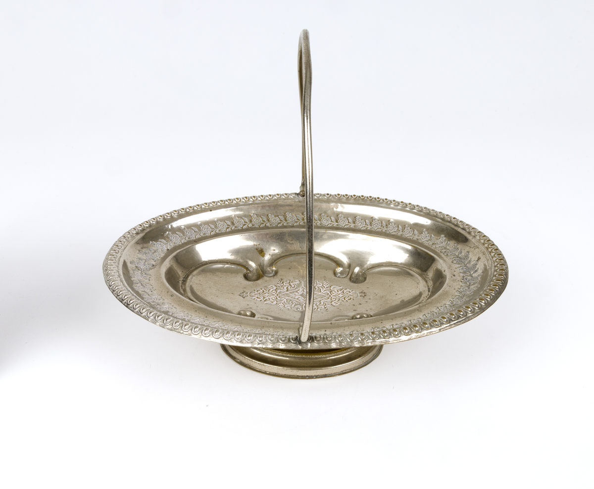 Skål. Oval og flat skål i form av en kurv, med håndtak og fot. Tynn metalltrådhåndtak fast på langsidene. Gravert dekor med plantemotiver med blomster og blader. Akantusblader som dekor midt i fatet.