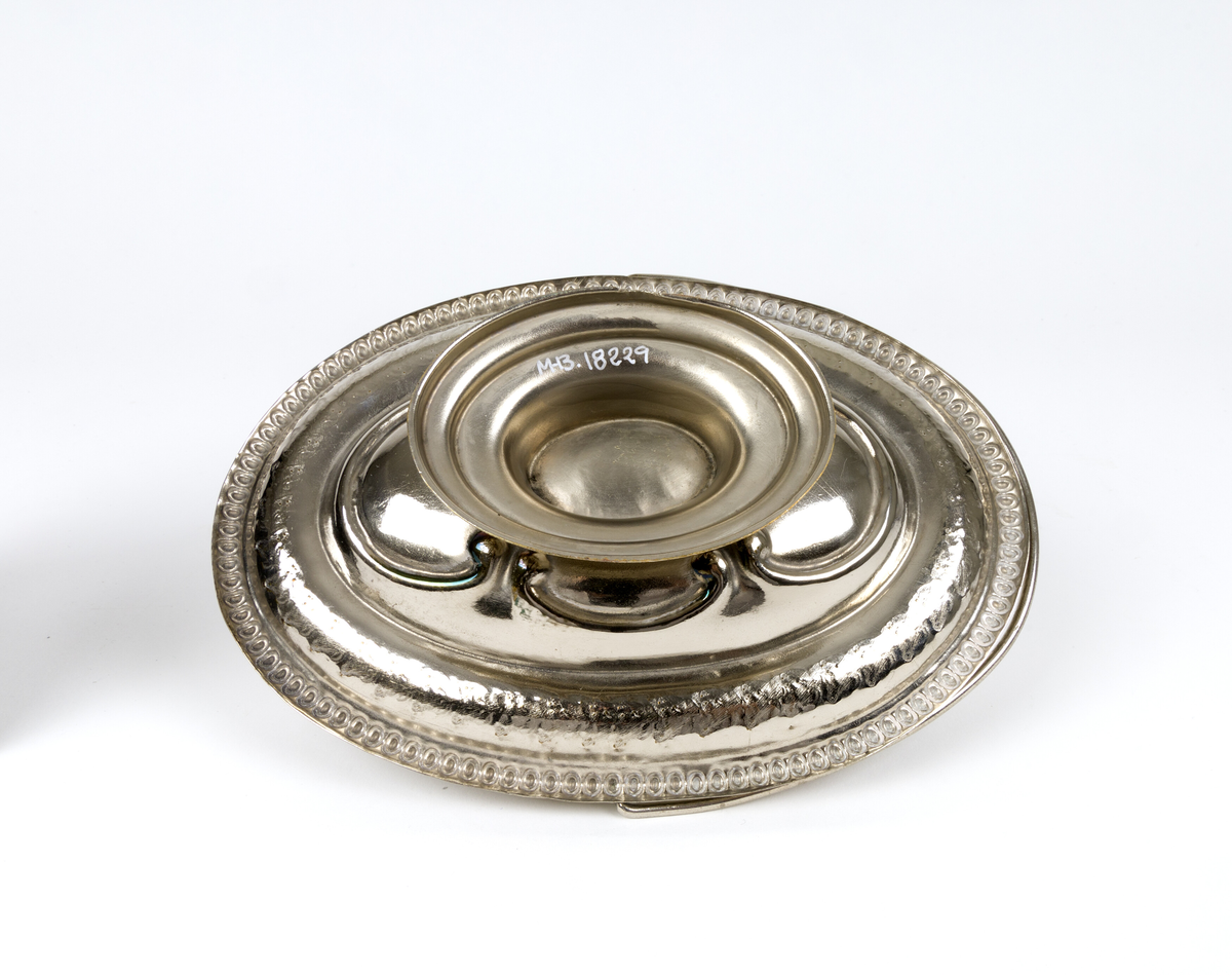 Skål. Oval og flat skål i form av en kurv, med håndtak og fot. Tynn metalltrådhåndtak fast på langsidene. Gravert dekor med plantemotiver med blomster og blader. Akantusblader som dekor midt i fatet.
