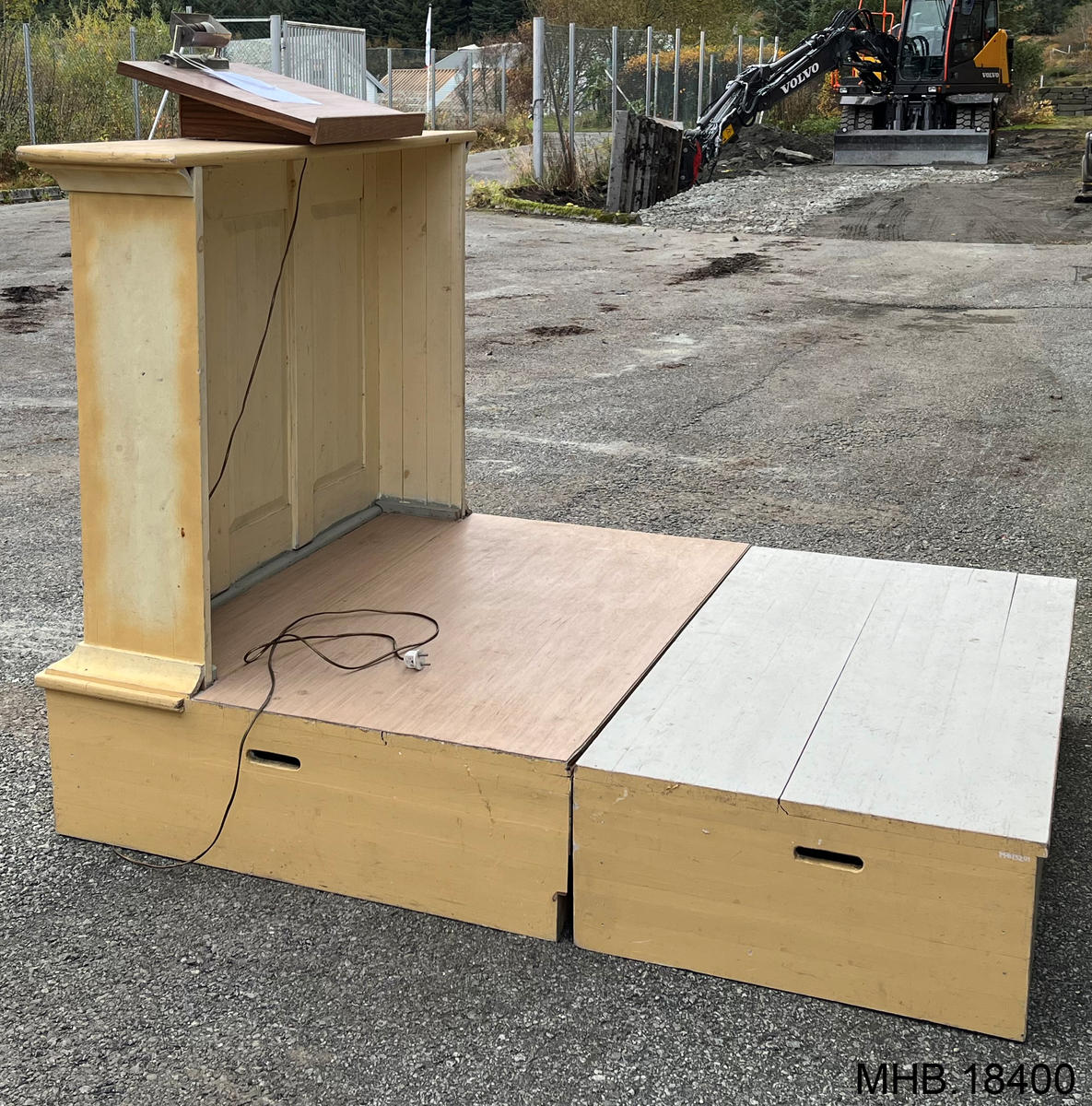 Prekestol av tre, montert på hevet platting. Tilhørende løs del til å forstørre platitngen. Håndtak på sidene av begge delene for flytting. Hylle til papirer med lys over motert på talerstolen. Trekors på forsiden.