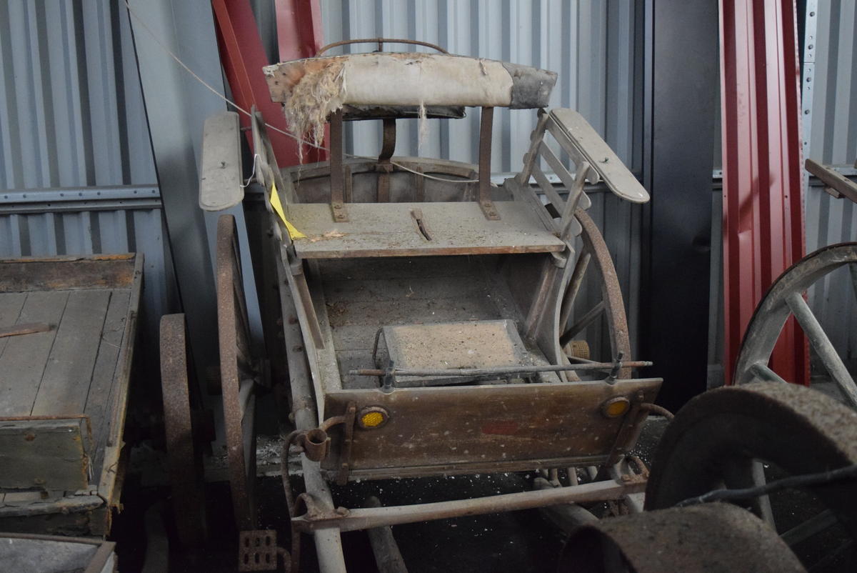 Kjerre med to hjul på aksling. Hjul av tre med 14(?) eiker og jernskoning. Benk med polstret rygg. Krakk foran. Polstert sete bak benken med lav metallkant bak og på sidene. Trebrett for skjerming mot stein og støv fremme.
