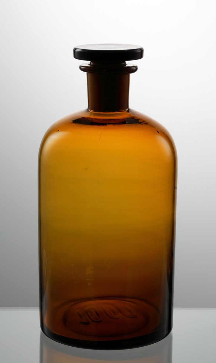 Apoteksflaska med glaspropp. Gulbrunt klarglas. Cylindrisk kropp med rundat axelparti. 

Inskrivet i huvudkatalogen 1972.