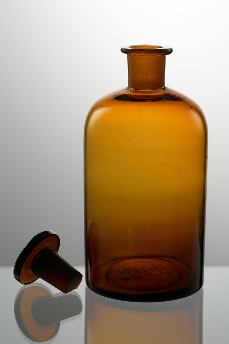 Apoteksflaska med glaspropp. Gulbrunt klarglas. Cylindrisk kropp med rundat axelparti. 

Inskrivet i huvudkatalogen 1972.