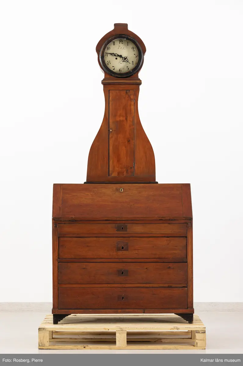 KLM 11514. Golvur. Snedklaffbyrå med klockfodral och urverk. Av furu betsat i brunt med svartmålade detaljer. Nedre delen av golvuret i form av en snedklaffbyrå med fyra lådor samt snedklaff, inuti med smålådor. Klockfodralet är fiolformat med dörr mitt på. Rund öppning för urtavlan, huvens glas saknas. Urtavla och visare av metall. Urtavlan målad i vitt med svarta siffror och signerad i svart: A.A.S. Mora. Urverk av stål och trä, tillhörande nyckel finns. Pendel med rund platta av mässing i form av en rocaille. Lod saknas.