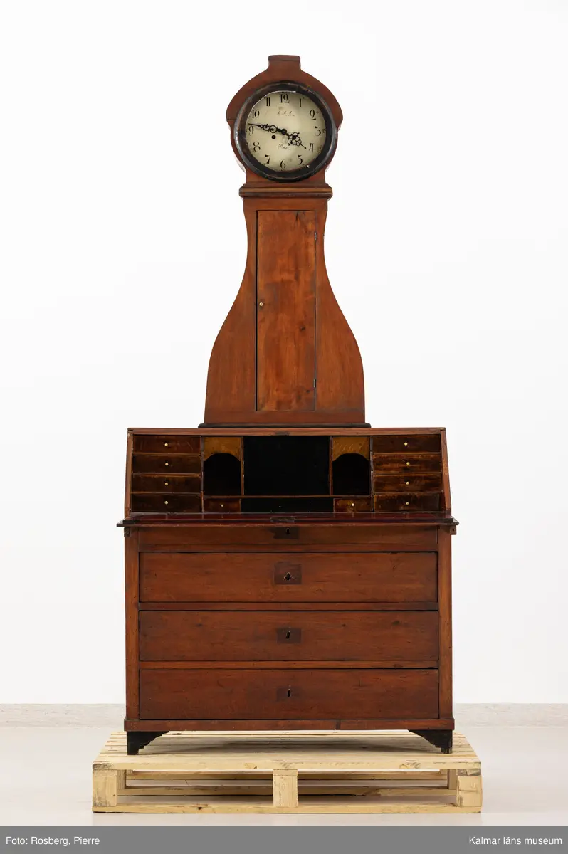 KLM 11514. Golvur. Snedklaffbyrå med klockfodral och urverk. Av furu betsat i brunt med svartmålade detaljer. Nedre delen av golvuret i form av en snedklaffbyrå med fyra lådor samt snedklaff, inuti med smålådor. Klockfodralet är fiolformat med dörr mitt på. Rund öppning för urtavlan, huvens glas saknas. Urtavla och visare av metall. Urtavlan målad i vitt med svarta siffror och signerad i svart: A.A.S. Mora. Urverk av stål och trä, tillhörande nyckel finns. Pendel med rund platta av mässing i form av en rocaille. Lod saknas.