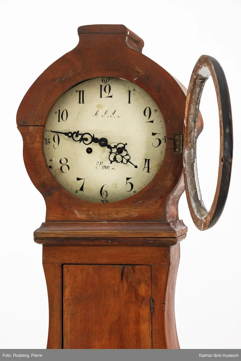 KLM 11514. Golvur. Snedklaffbyrå med klockfodral och urverk. Av furu betsat i brunt med svartmålade detaljer. Nedre delen av golvuret i form av en snedklaffbyrå med fyra lådor samt snedklaff, inuti med smålådor. Klockfodralet är fiolformat med dörr mitt på. Rund öppning för urtavlan, huvens glas saknas. Urtavla och visare av metall. Urtavlan målad i vitt med svarta siffror och signerad i svart: A.A.S. Mora. Urverk av stål och trä, tillhörande nyckel finns. Pendel med rund platta av mässing i form av en rocaille. Lod saknas.