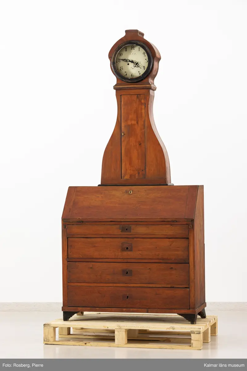 KLM 11514. Golvur. Snedklaffbyrå med klockfodral och urverk. Av furu betsat i brunt med svartmålade detaljer. Nedre delen av golvuret i form av en snedklaffbyrå med fyra lådor samt snedklaff, inuti med smålådor. Klockfodralet är fiolformat med dörr mitt på. Rund öppning för urtavlan, huvens glas saknas. Urtavla och visare av metall. Urtavlan målad i vitt med svarta siffror och signerad i svart: A.A.S. Mora. Urverk av stål och trä, tillhörande nyckel finns. Pendel med rund platta av mässing i form av en rocaille. Lod saknas.