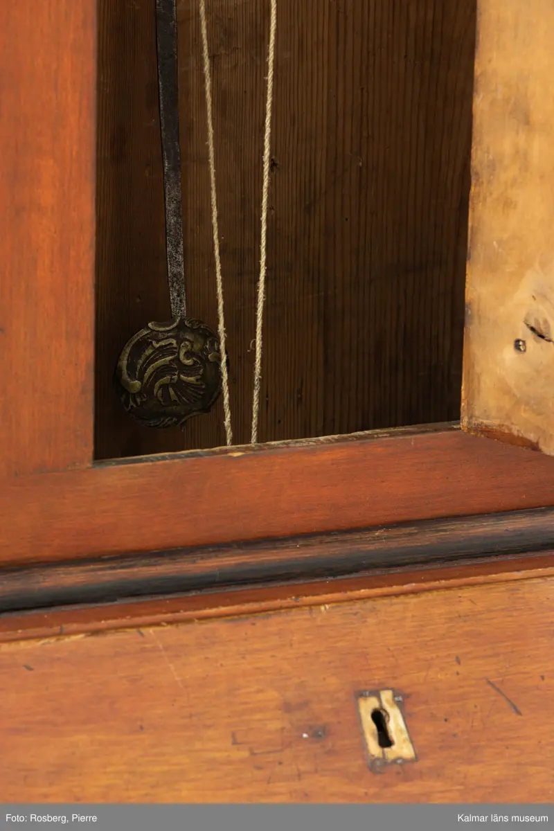 KLM 11514. Golvur. Snedklaffbyrå med klockfodral och urverk. Av furu betsat i brunt med svartmålade detaljer. Nedre delen av golvuret i form av en snedklaffbyrå med fyra lådor samt snedklaff, inuti med smålådor. Klockfodralet är fiolformat med dörr mitt på. Rund öppning för urtavlan, huvens glas saknas. Urtavla och visare av metall. Urtavlan målad i vitt med svarta siffror och signerad i svart: A.A.S. Mora. Urverk av stål och trä, tillhörande nyckel finns. Pendel med rund platta av mässing i form av en rocaille. Lod saknas.