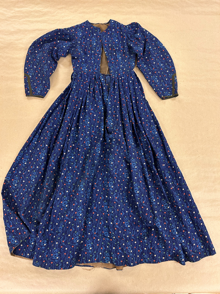 Klänning i blå bottenfärg, vattrat mönster, prickar i gult, rött och grönt. Slätt figurskuret liv med insydda fjädrar. Valben i ryggsöm och insnitt fram. Knäppning framtill med 13 st hyskor och hakar, varav flera saknas. Ingen krage. Isydd ärm med lagda veck över axeln. Passpoal i sömmen. Smalt markerat ryggstycke med svarta passpoalmarkerade sömmar. En vid ärm som smalnar vid handleden, vilken är kantad med svarta bomullsband, vilka nu är gröna. Kjolen har lagda veck runt om, sprund mitt fram. Sydd av 4 hela våder och en kilskuren våd. Sparkband av samma bruna linnelärft som livets foder. Vänster ärm är fodrad med brunt kattuntyg med mönster av vita prickar på brun botten, däröver brandgula blommor och streckliknande mönster. Klänningen är lagad på flera ställen och hålig.