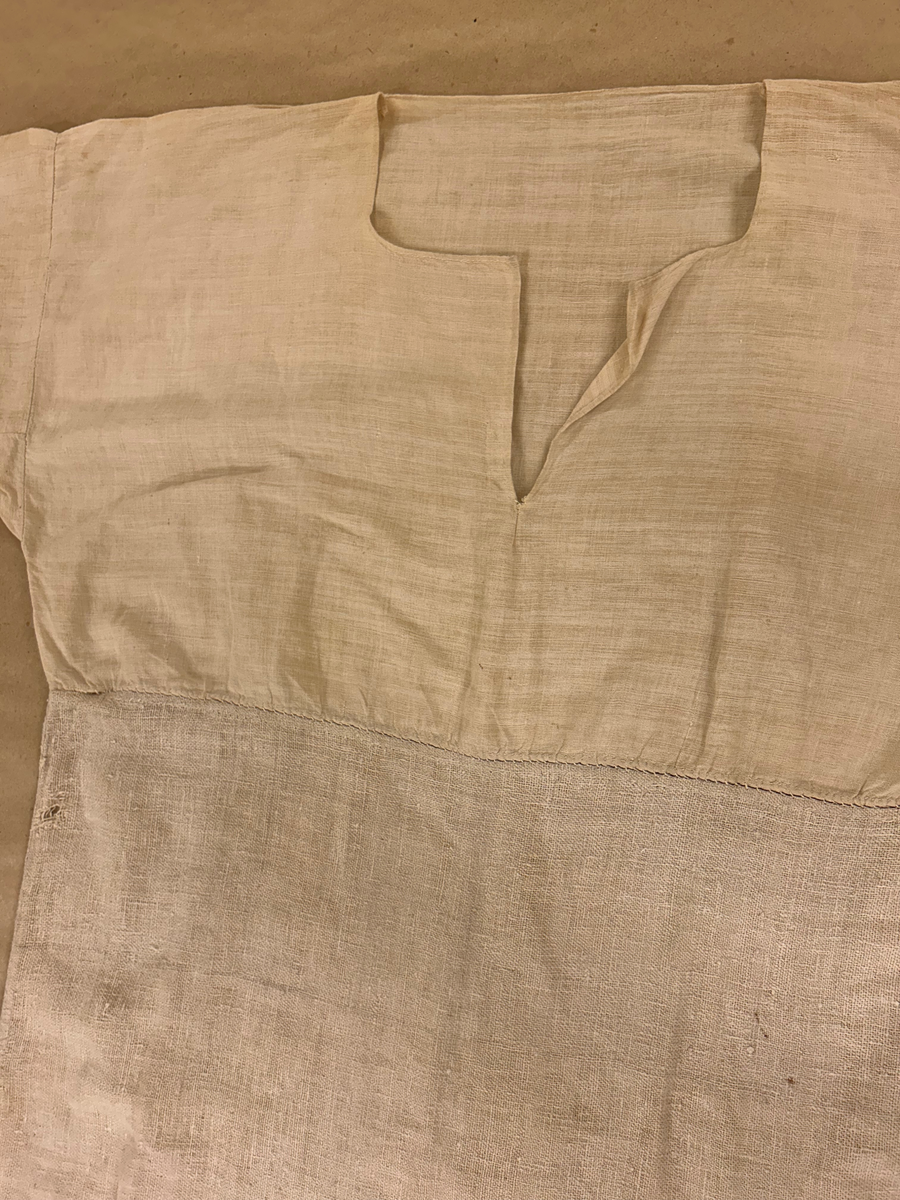 Särk i vitt, tuskaftat linne. Livet skuret i ett rakt stycke, ingen axelsöm. Sprund framtill, rundad halsringning. Rakt skuren ärm med spjäll. Nedre delen av grövre lingarn påsydd på livet strax neranför sprundet. Isydda kilar i sidsömmarna.