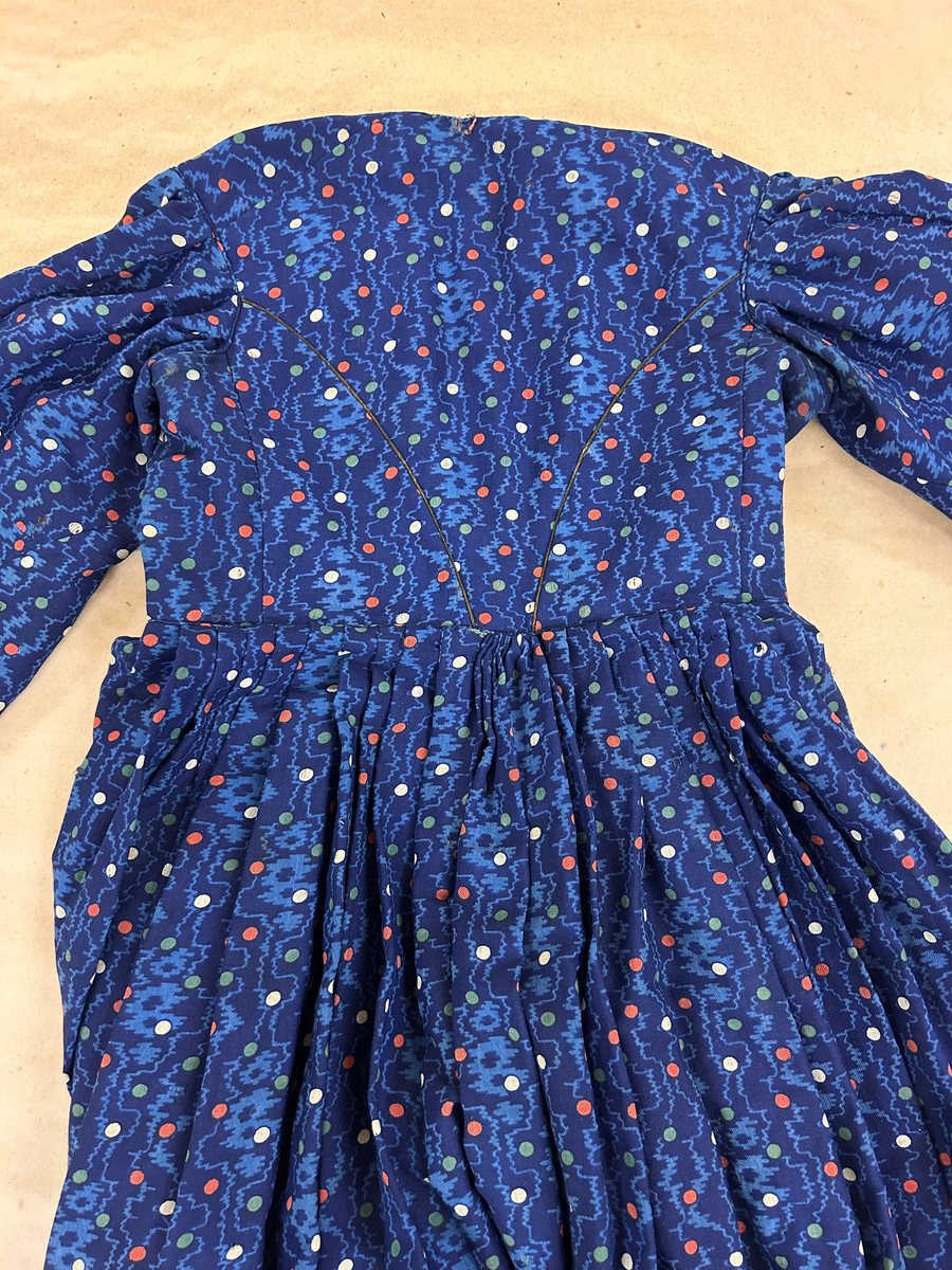 Klänning i blå bottenfärg, vattrat mönster, prickar i gult, rött och grönt. Slätt figurskuret liv med insydda fjädrar. Valben i ryggsöm och insnitt fram. Knäppning framtill med 13 st hyskor och hakar, varav flera saknas. Ingen krage. Isydd ärm med lagda veck över axeln. Passpoal i sömmen. Smalt markerat ryggstycke med svarta passpoalmarkerade sömmar. En vid ärm som smalnar vid handleden, vilken är kantad med svarta bomullsband, vilka nu är gröna. Kjolen har lagda veck runt om, sprund mitt fram. Sydd av 4 hela våder och en kilskuren våd. Sparkband av samma bruna linnelärft som livets foder. Vänster ärm är fodrad med brunt kattuntyg med mönster av vita prickar på brun botten, däröver brandgula blommor och streckliknande mönster. Klänningen är lagad på flera ställen och hålig.