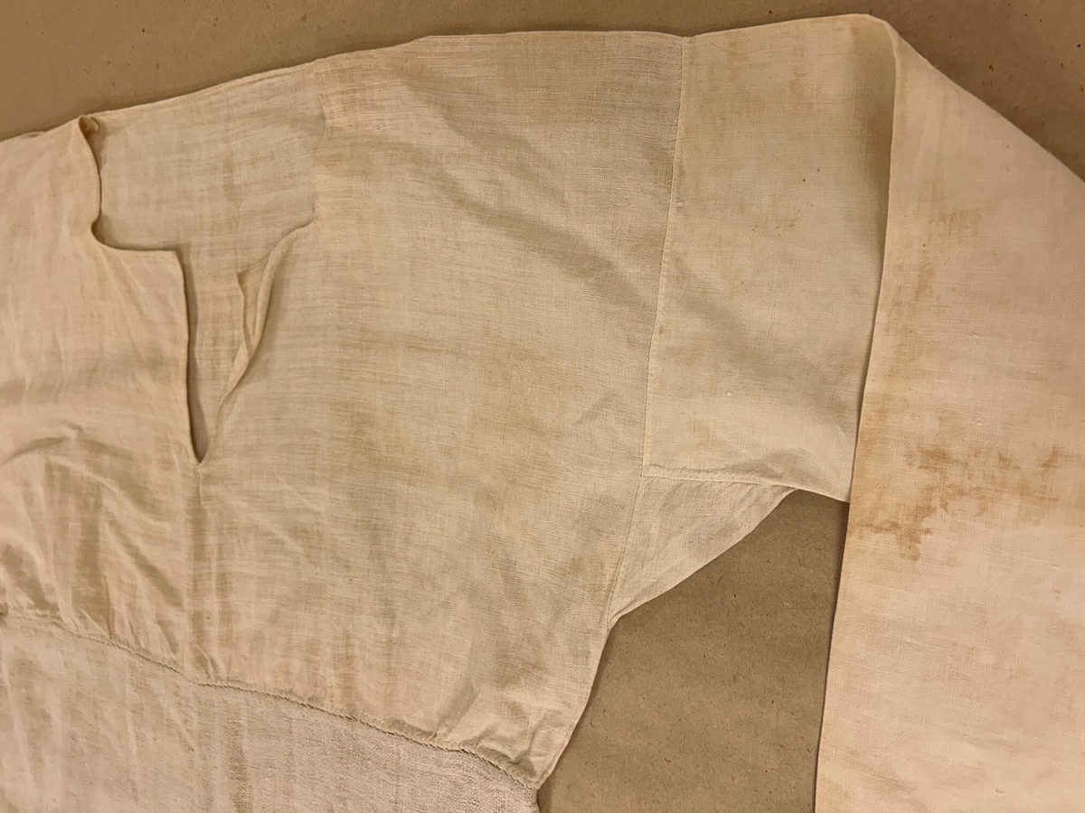 Särk i vitt, tuskaftat linne. Livet skuret i ett rakt stycke, ingen axelsöm. Sprund framtill, rundad halsringning. Rakt skuren ärm med spjäll. Nedre delen av grövre lingarn påsydd på livet strax neranför sprundet. Isydda kilar i sidsömmarna.