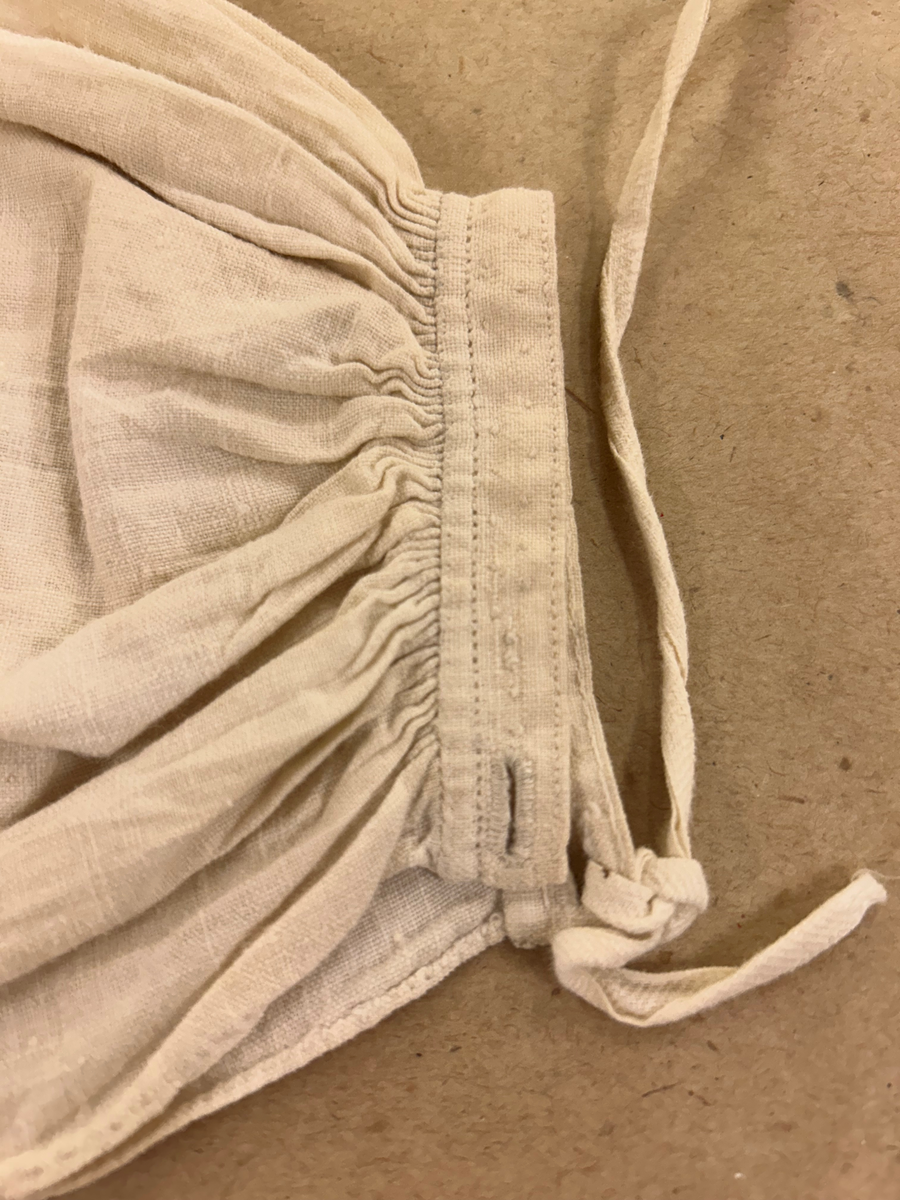 Särk av tuskaftat linne. Fram- och bakstycke av ett sammanhängande stycke isydda kilar i sidosömmen. Förstärkning över axlarna, sprund framtil. Rundad halsringning. Rak ärm med spjäll, rynkad mot axeln och handlinning. Tränsade knapphål och sydda prickstickstygn på linningen, knytband.