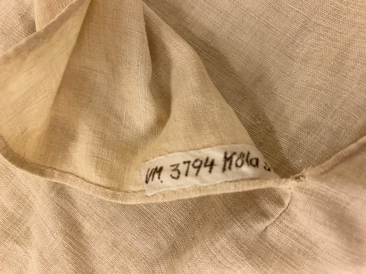 Särk i vitt, tuskaftat linne. Livet skuret i ett rakt stycke, ingen axelsöm. Sprund framtill, rundad halsringning. Rakt skuren ärm med spjäll. Nedre delen av grövre lingarn påsydd på livet strax neranför sprundet. Isydda kilar i sidsömmarna.