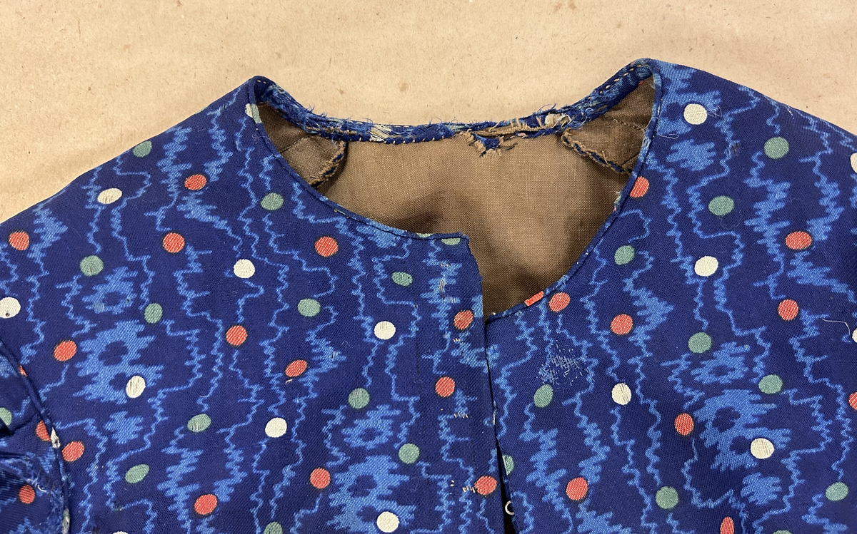 Klänning i blå bottenfärg, vattrat mönster, prickar i gult, rött och grönt. Slätt figurskuret liv med insydda fjädrar. Valben i ryggsöm och insnitt fram. Knäppning framtill med 13 st hyskor och hakar, varav flera saknas. Ingen krage. Isydd ärm med lagda veck över axeln. Passpoal i sömmen. Smalt markerat ryggstycke med svarta passpoalmarkerade sömmar. En vid ärm som smalnar vid handleden, vilken är kantad med svarta bomullsband, vilka nu är gröna. Kjolen har lagda veck runt om, sprund mitt fram. Sydd av 4 hela våder och en kilskuren våd. Sparkband av samma bruna linnelärft som livets foder. Vänster ärm är fodrad med brunt kattuntyg med mönster av vita prickar på brun botten, däröver brandgula blommor och streckliknande mönster. Klänningen är lagad på flera ställen och hålig.