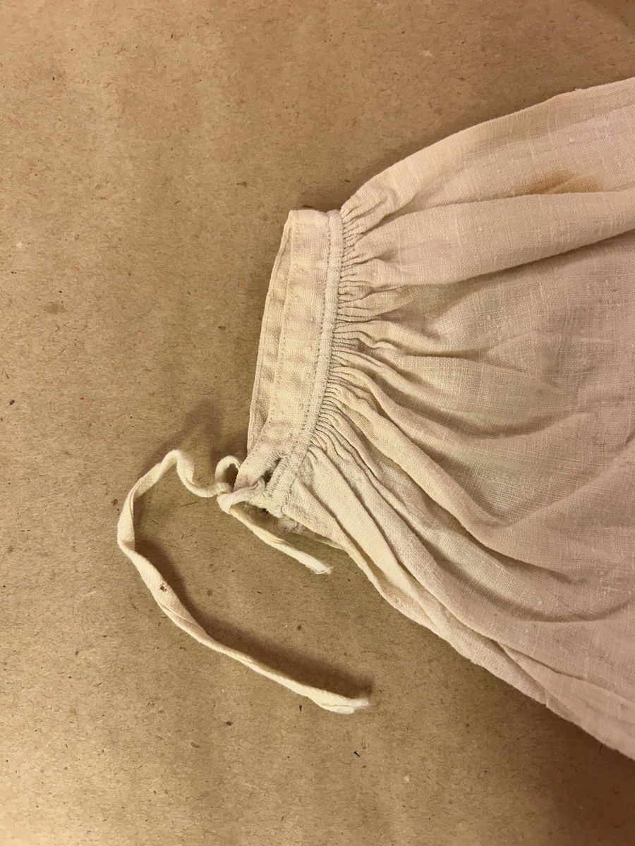 Särk av tuskaftat linne. Fram- och bakstycke av ett sammanhängande stycke isydda kilar i sidosömmen. Förstärkning över axlarna, sprund framtil. Rundad halsringning. Rak ärm med spjäll, rynkad mot axeln och handlinning. Tränsade knapphål och sydda prickstickstygn på linningen, knytband.